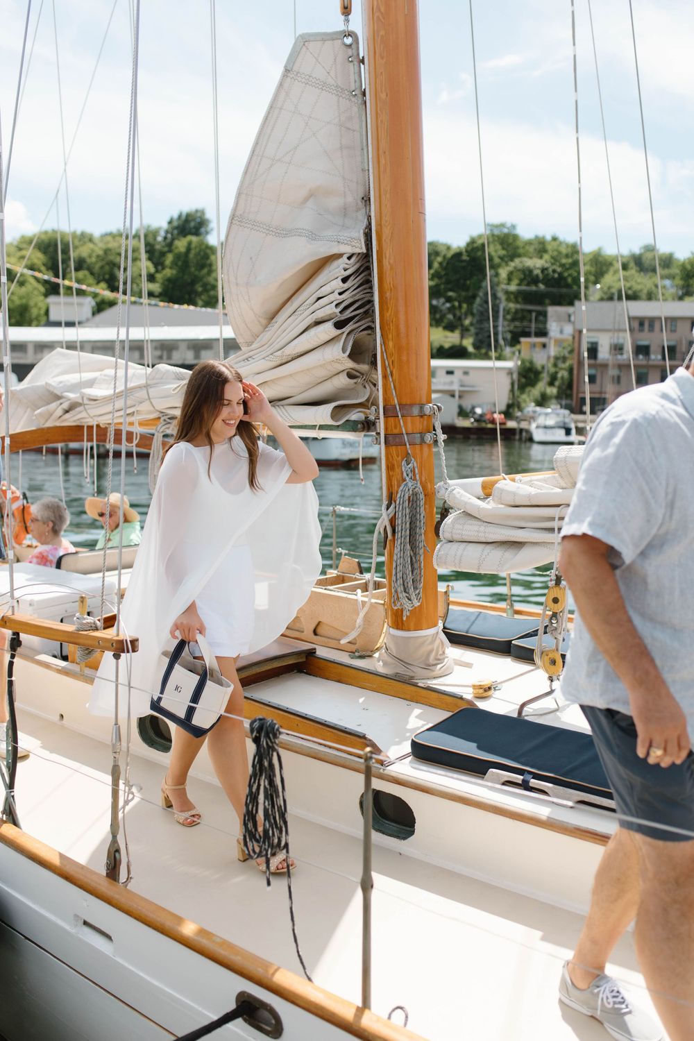 Woman in white dress boarding a classic wooden sailboat at a sunny marina on a beautiful day.