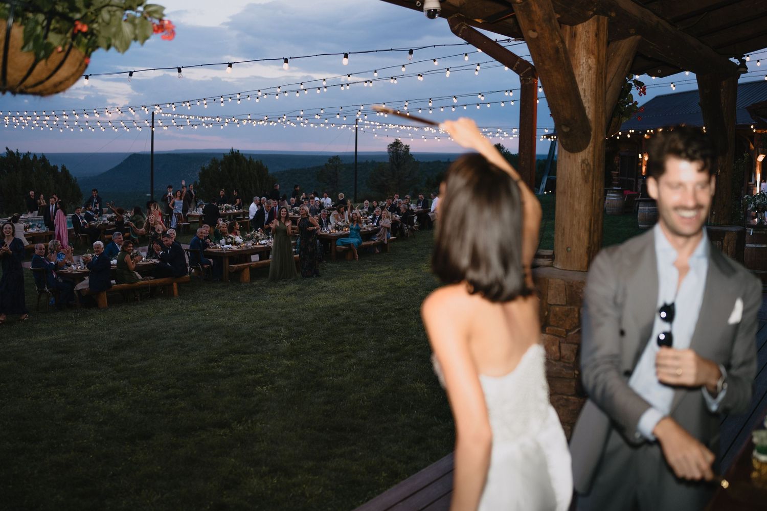 Bride and groom celebrate at outdoor wedding reception with string lights and mountain views at dusk.