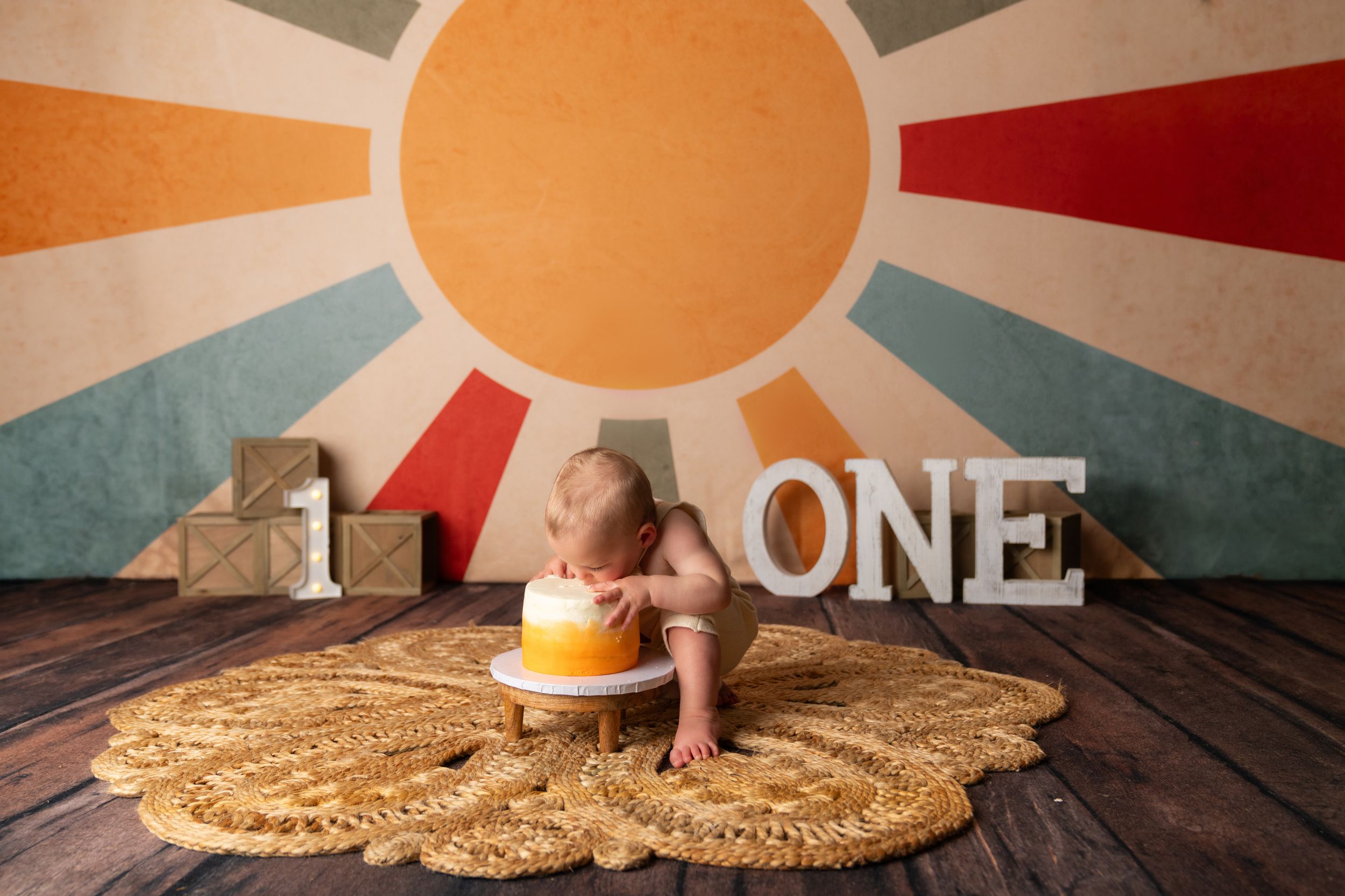 Baby smashing a yellow cake during a first birthday photoshoot with a colorful sun backdrop and ONE sign.