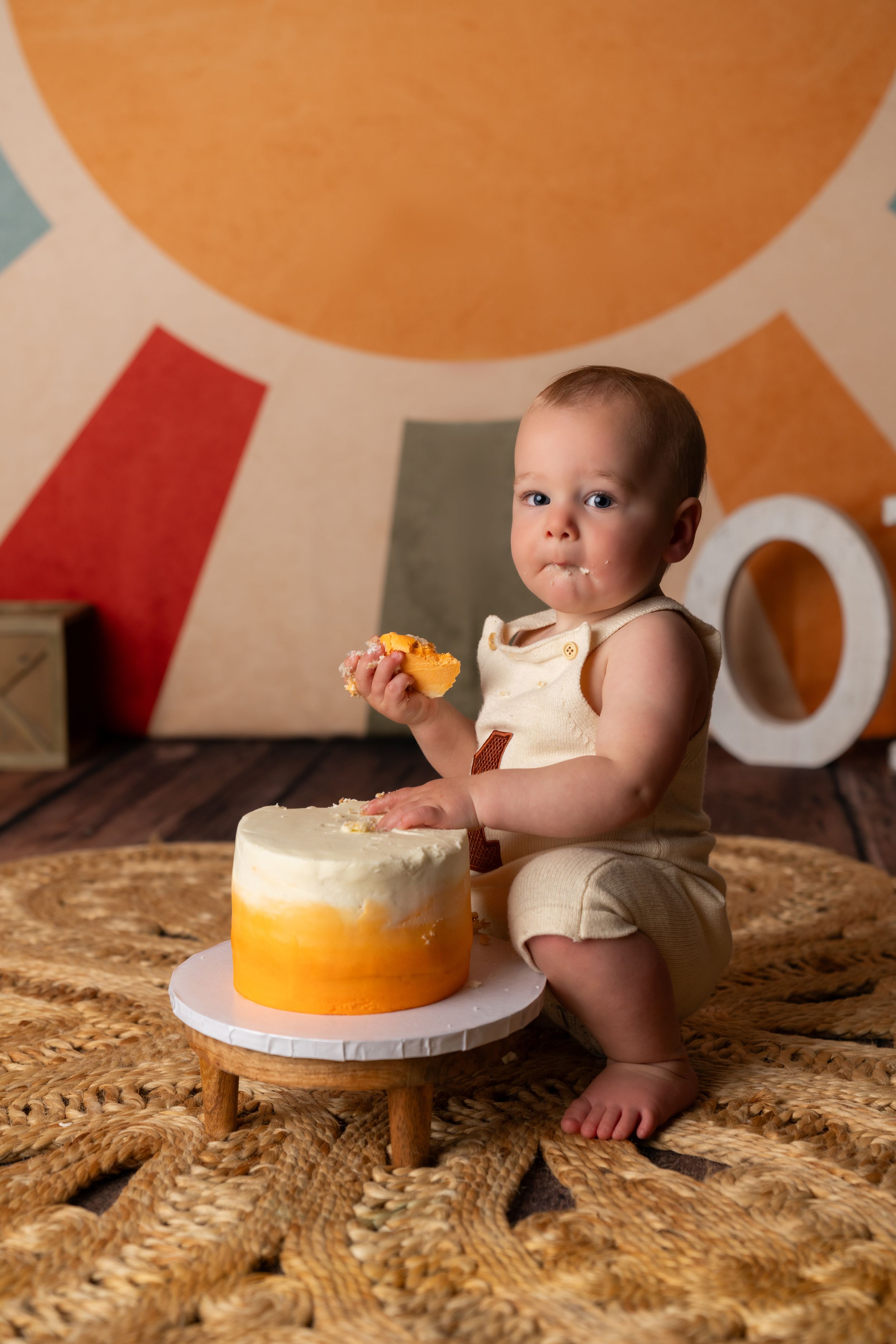 Baby in cream romper smashing first birthday cake with orange ombre frosting on woven rug with colorful backdrop.