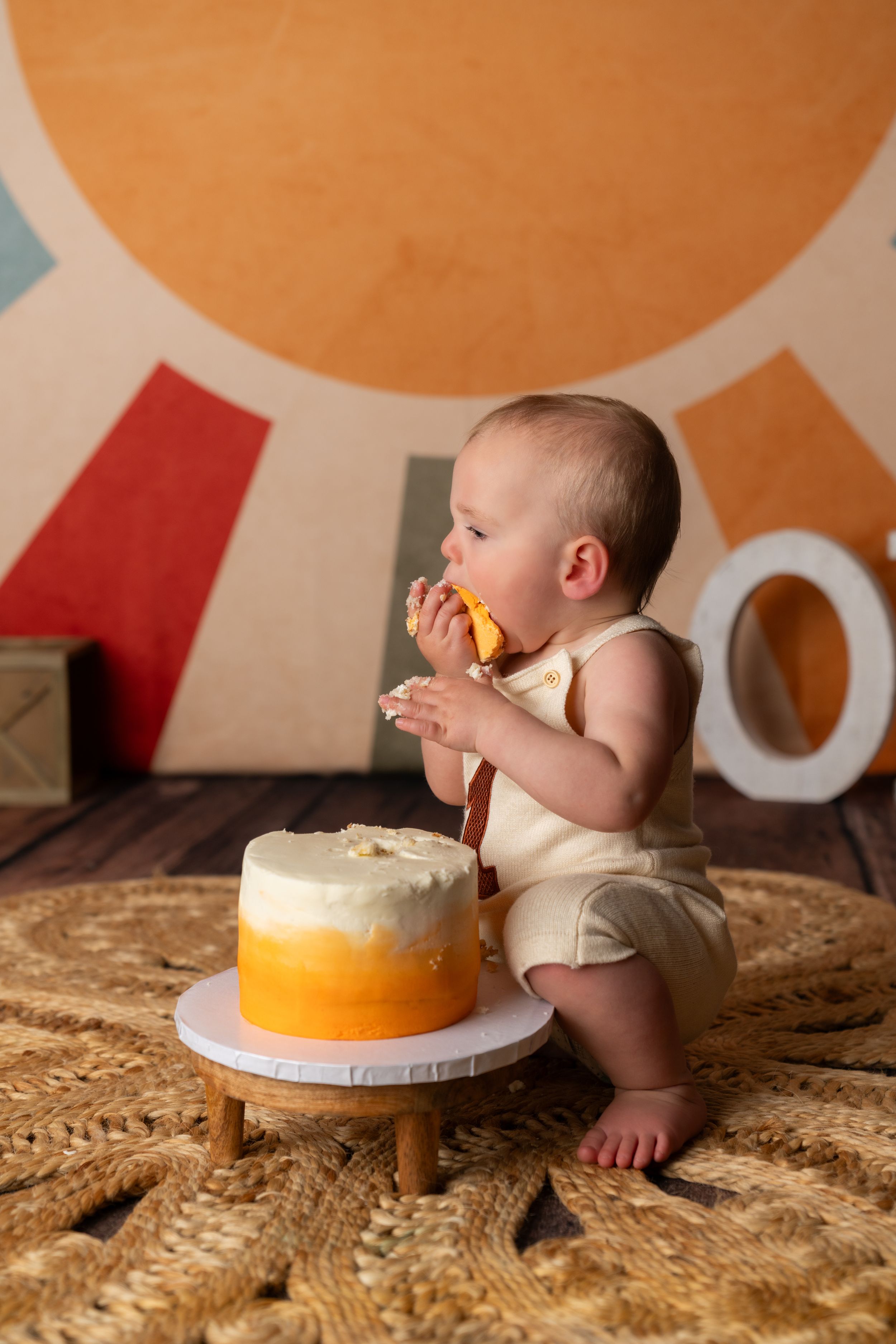 Baby in cream romper eating smash cake with orange ombre frosting during first birthday photo session.