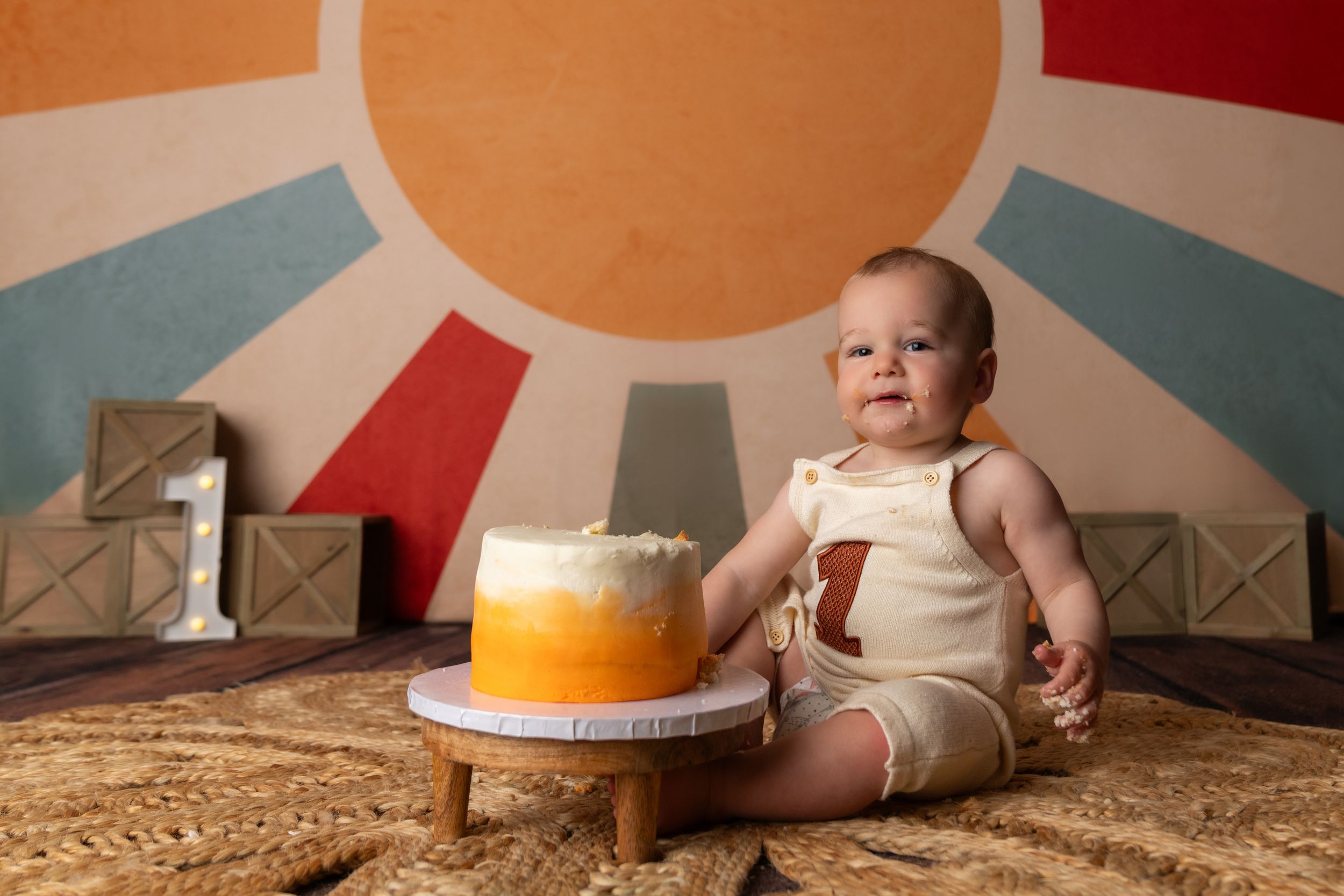 Baby in a onesie sits beside a yellow ombre smash cake in front of a colorful sun backdrop for a first birthday.