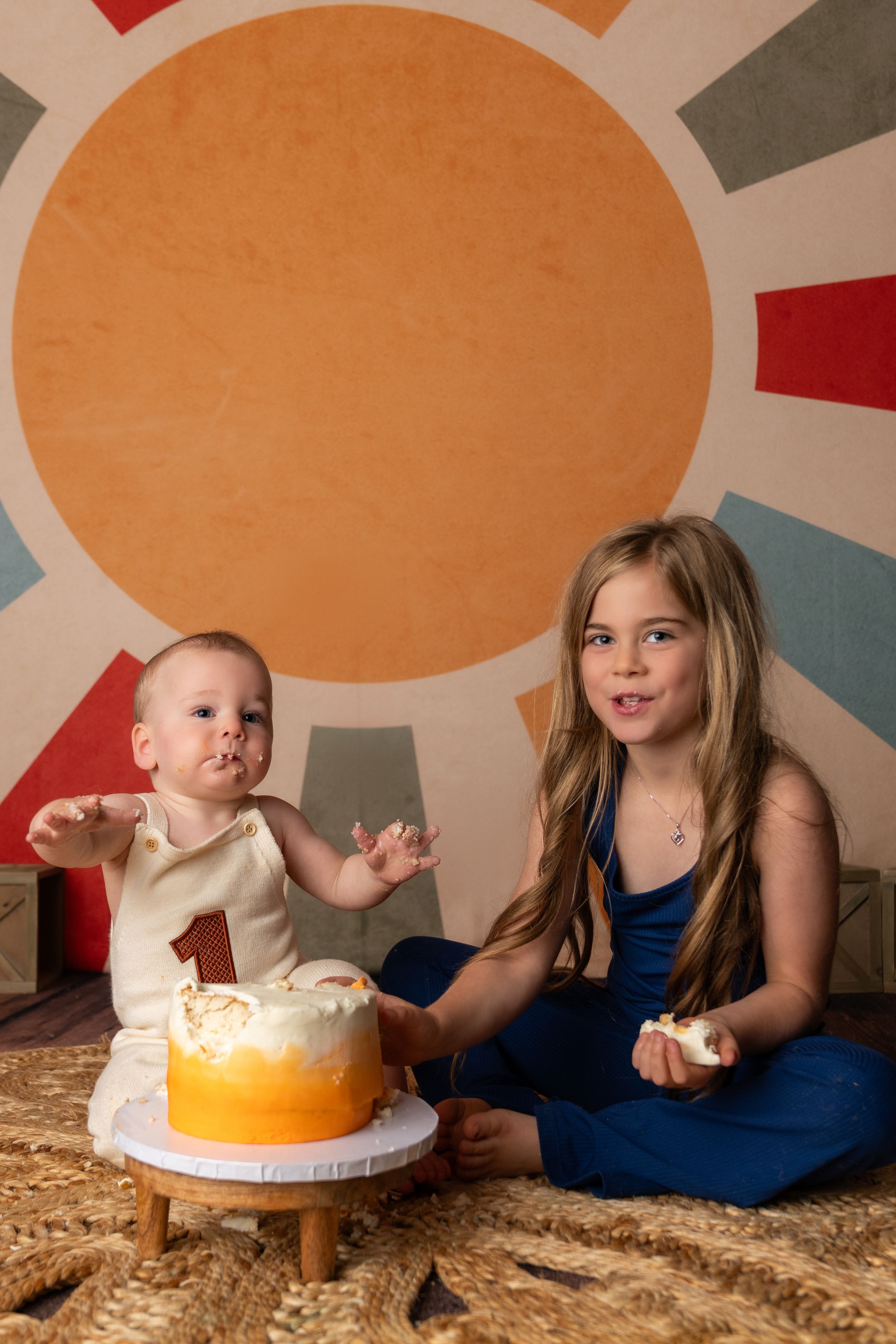 A baby celebrates their first birthday with an older sister beside a white cake with a number one candle on a colorful backdrop.
