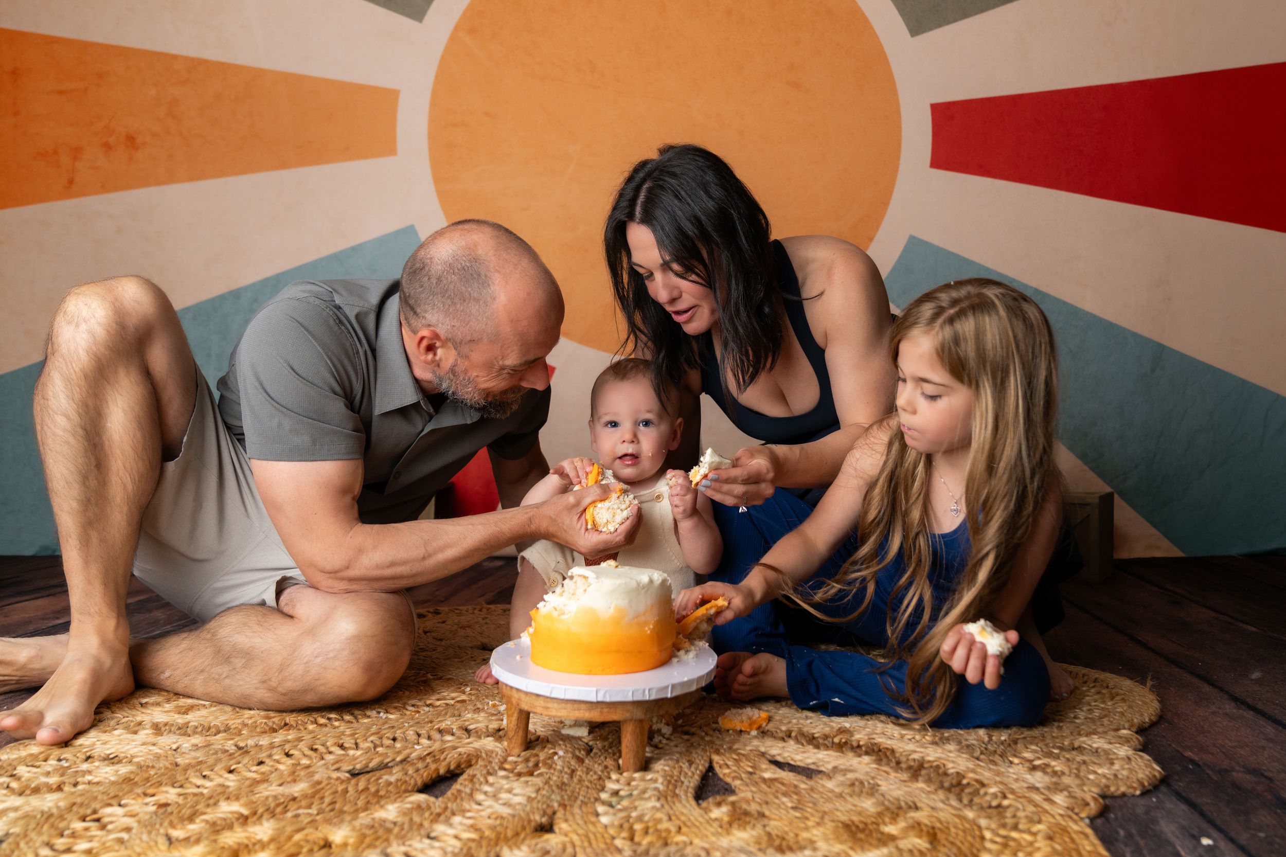 Family celebrating baby's first birthday with a smash cake, parents and sibling gathered on a rug.