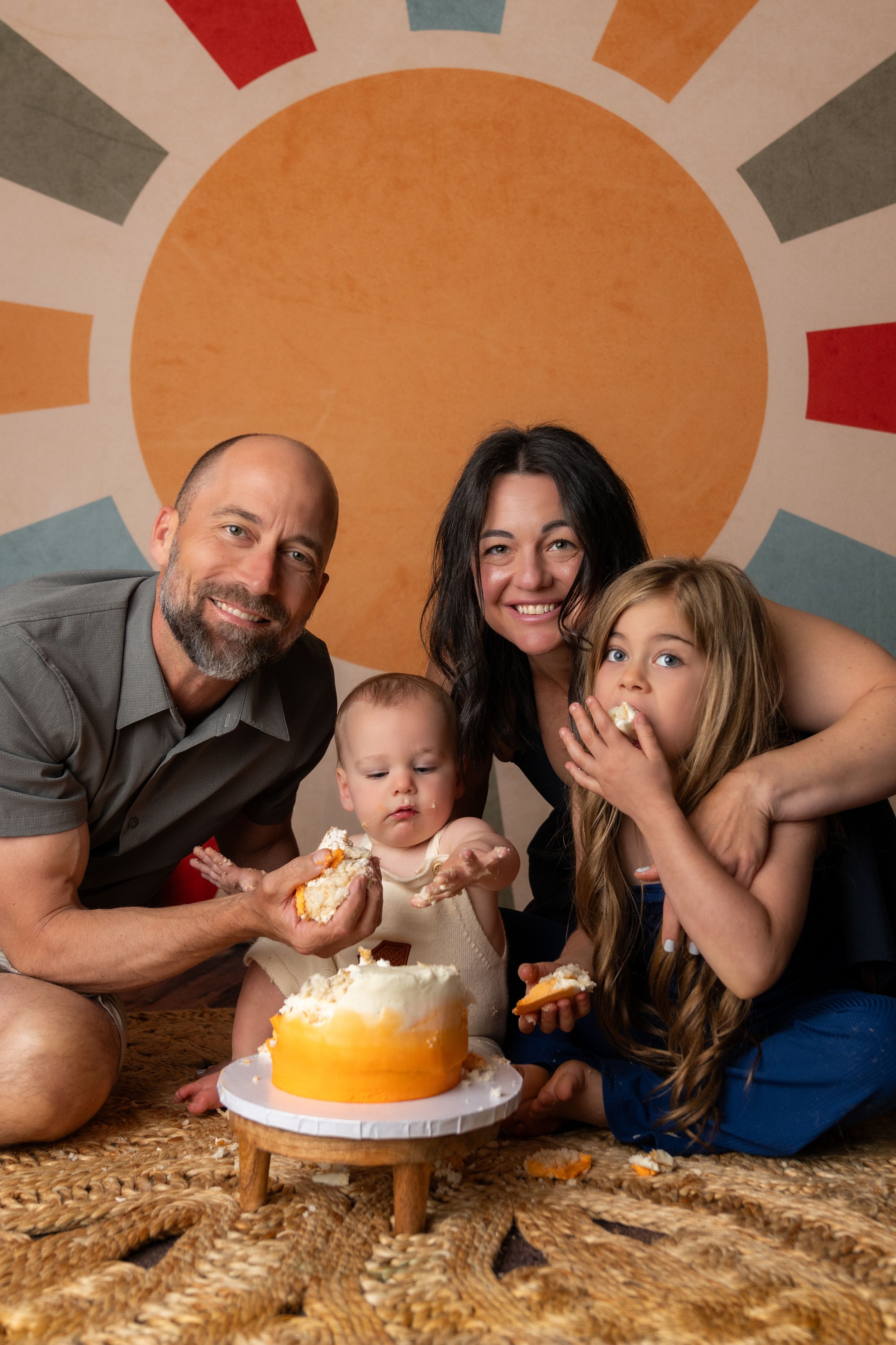 Family of four celebrating baby's first birthday cake smash with colorful sun backdrop and orange ombre cake.