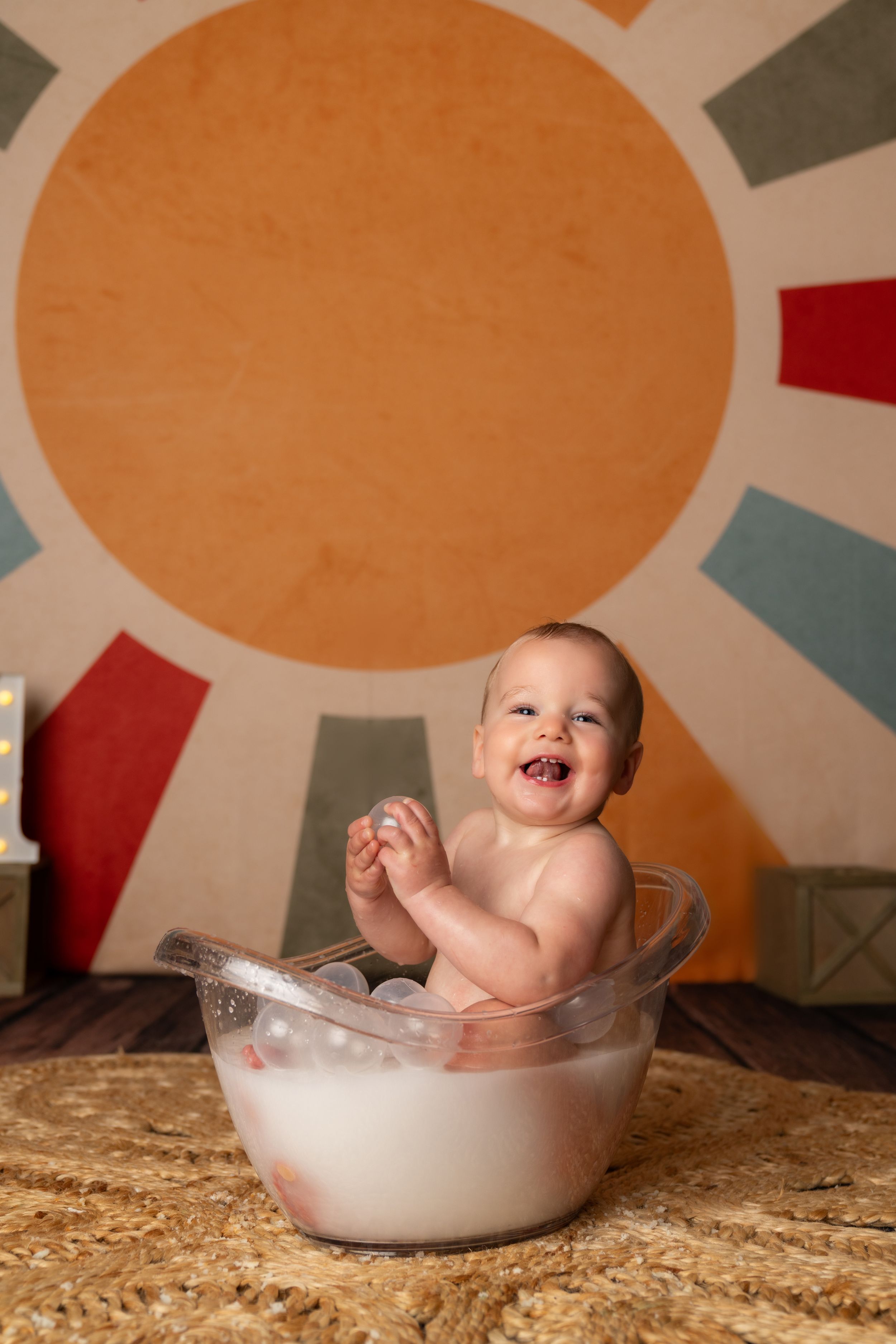 Happy baby sitting in a clear tub of milk, smiling and clapping, with a colorful sun mural backdrop.