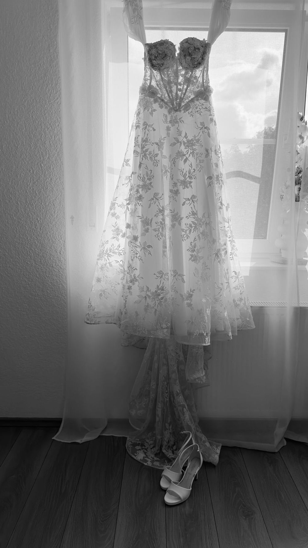 Elegant floral lace wedding gown hanging in window in black and white photo.