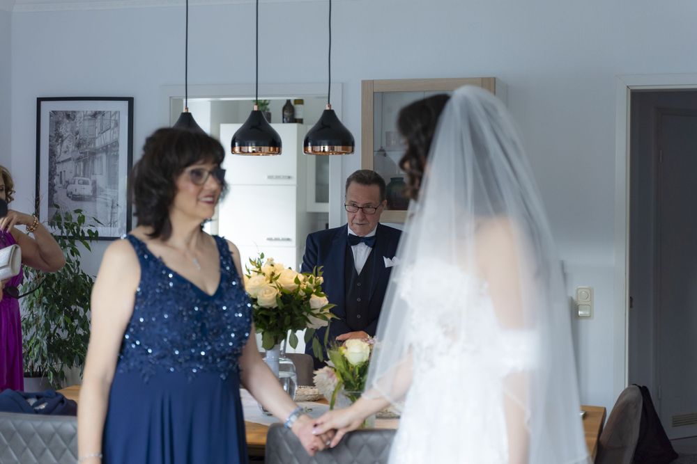 Bride in white gown and veil holds hands with woman in blue dress at wedding preparation.
