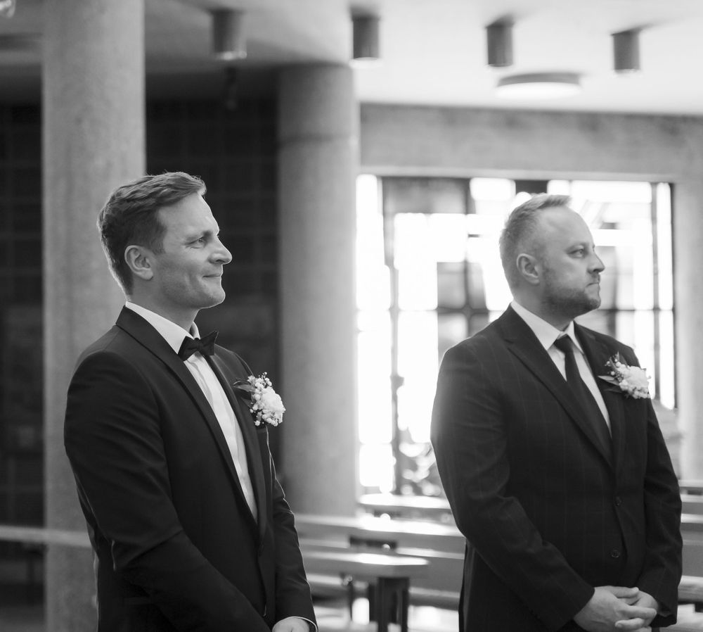 Two men in black tuxedos with boutonnieres standing at church altar in black and white.