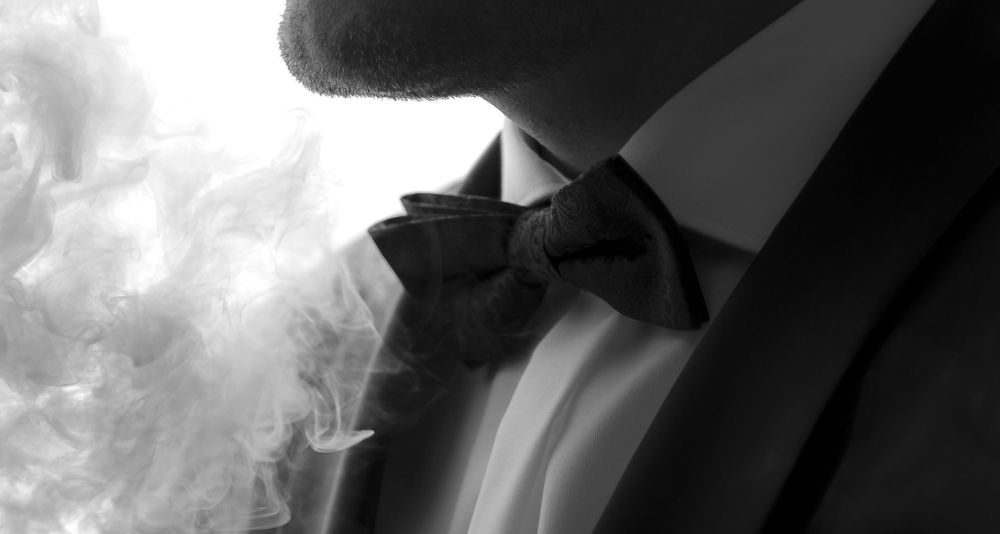 Elegant groom in black tuxedo and bow tie with smoke in black and white photo.