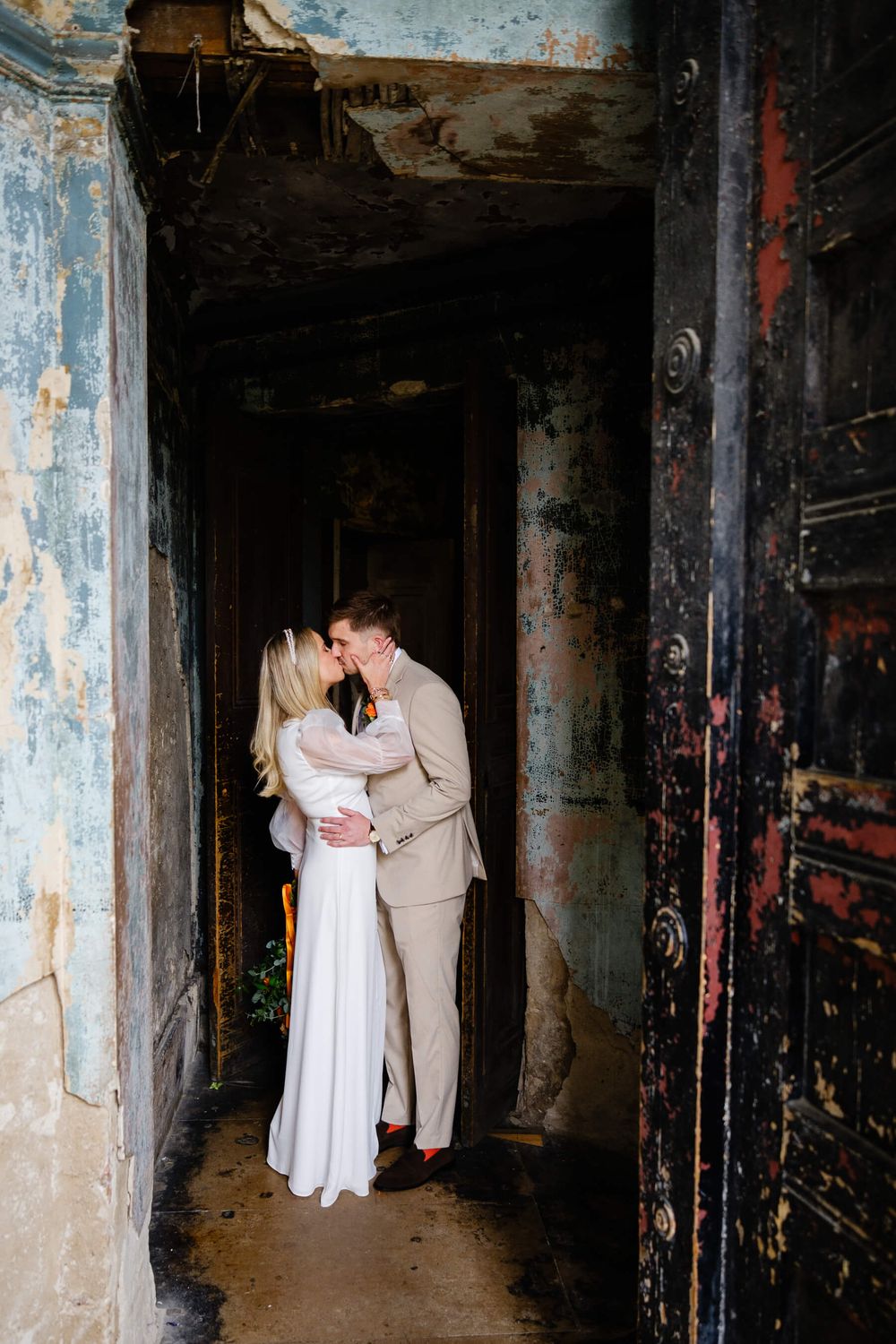 Bride and groom share a romantic kiss in a rustic, weathered doorway with peeling paint and aged walls.
