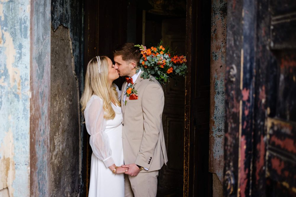 Bride and groom sharing a kiss in a rustic doorway, with colorful orange and red floral bouquet accents. at the asylum London