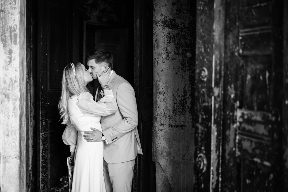 Black and white photo of a couple sharing a romantic kiss in a doorway, dressed in elegant wedding attire.