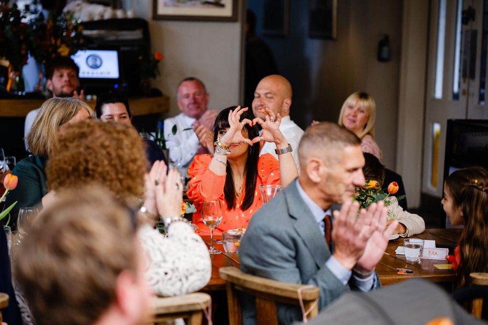 Wedding guests clapping and celebrating at a reception dinner, woman making heart shape with hands at decorated table.