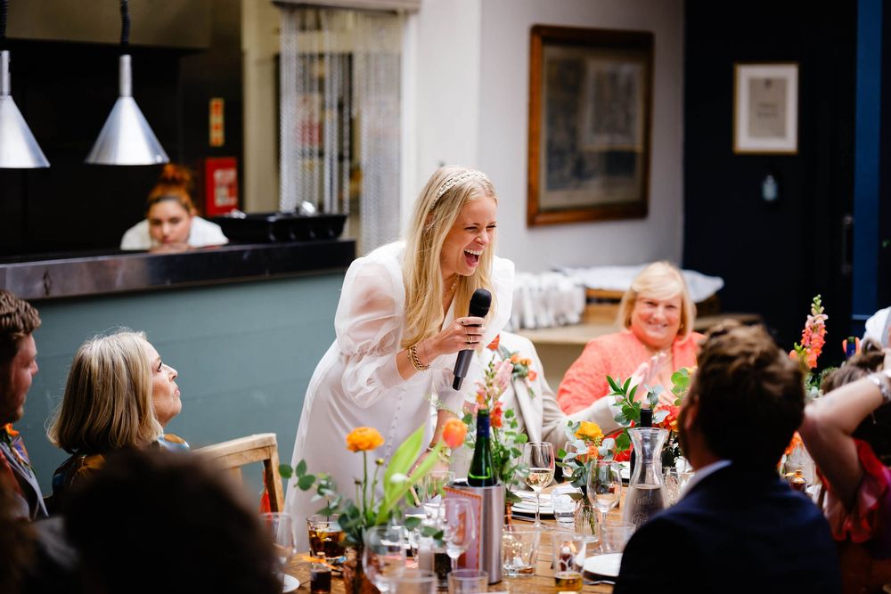 Bride giving a speech at wedding reception dinner, holding microphone, surrounded by guests and floral centerpieces.