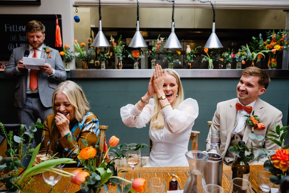 Joyful wedding reception guests laughing at speeches, with colorful floral arrangements and warm pendant lights.