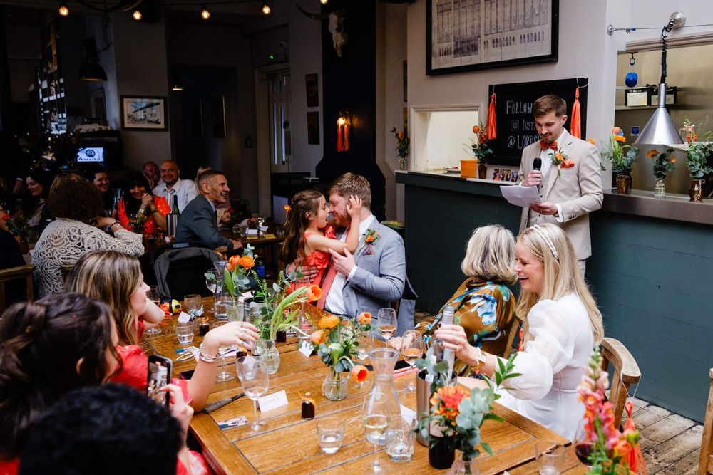 Wedding guests applaud as father and daughter kiss at rustic wooden table during indoor reception with colorful floral centerpieces.