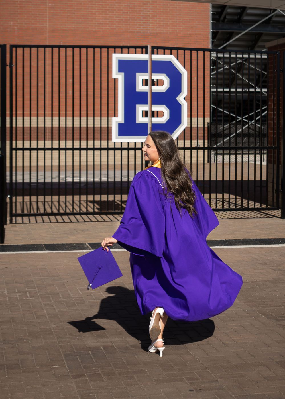 Graduate in purple cap and gown walks past a gate with a large blue letter B school logo.