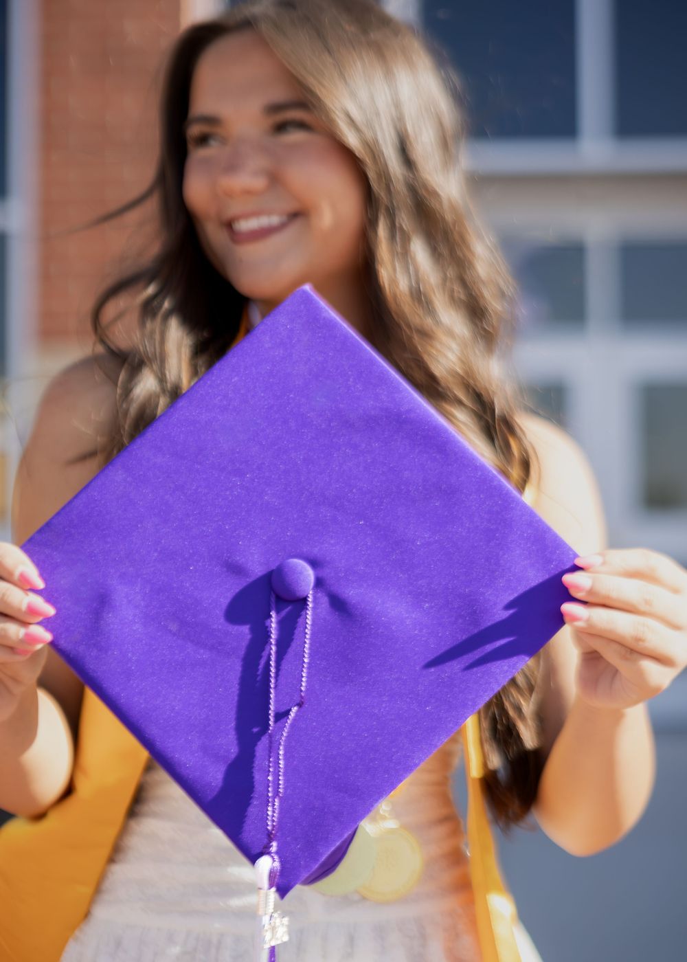 Smiling graduate holds up a purple mortarboard cap outdoors in front of a brick building.