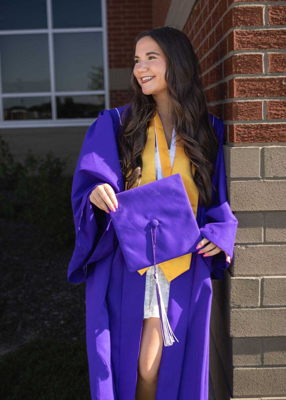 Brunette graduate in purple cap and gown smiles while leaning against a brick wall outdoors.