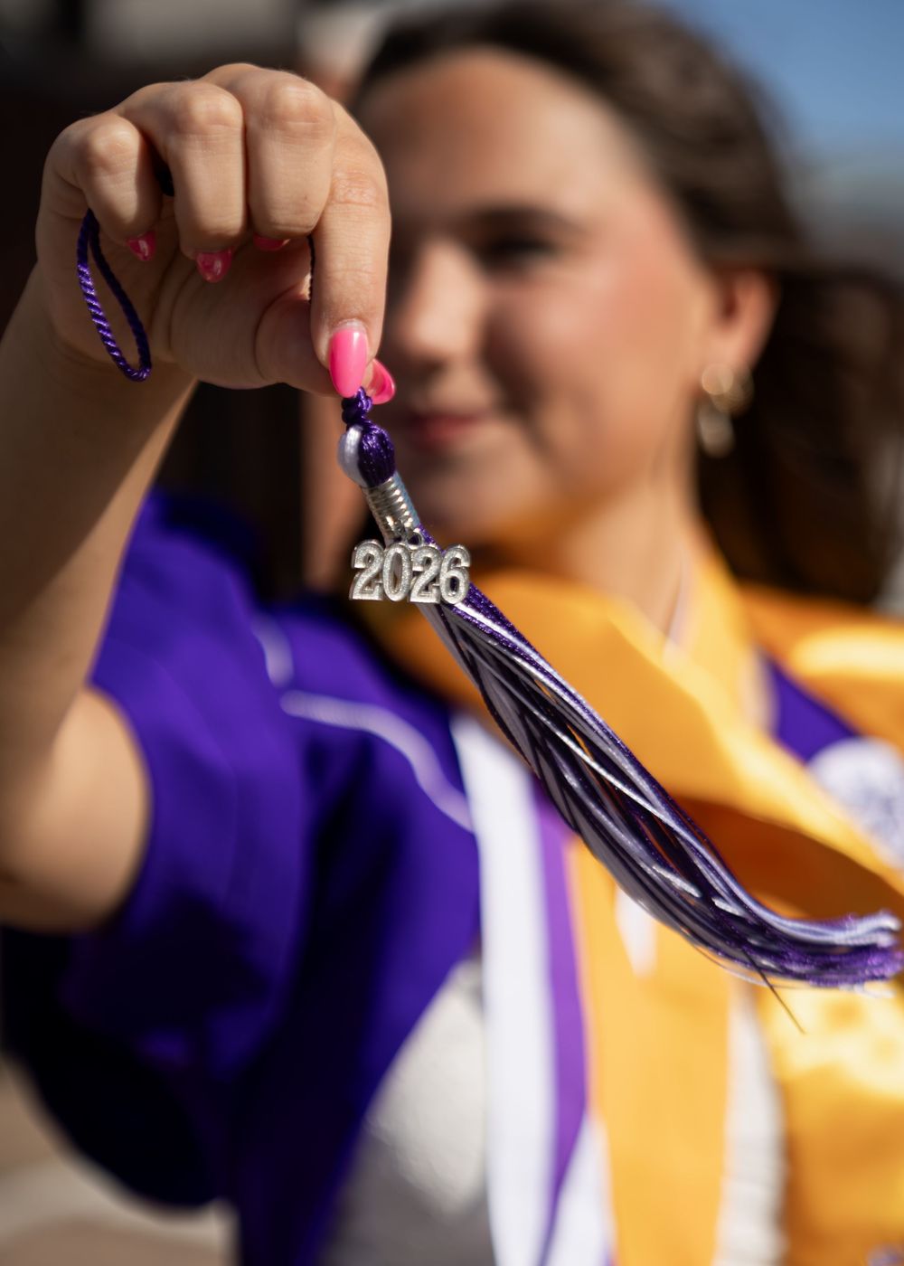 Close-up of a graduate holding a 2026 tassel with pink nails wearing a purple and gold graduation gown.