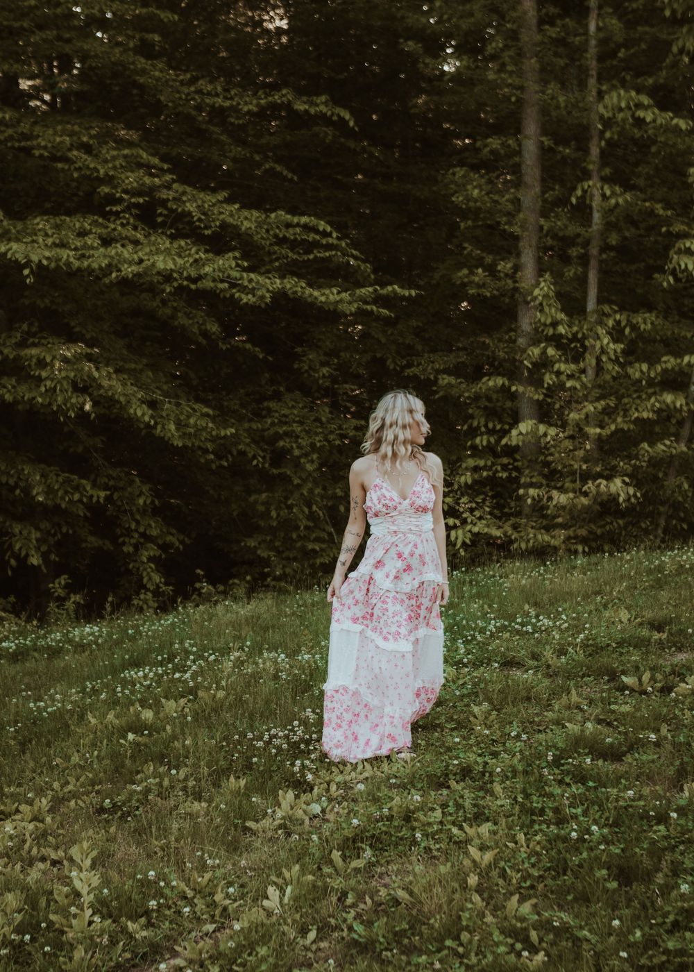 Blonde woman in a pink floral maxi dress stands in a green meadow surrounded by tall evergreen trees.