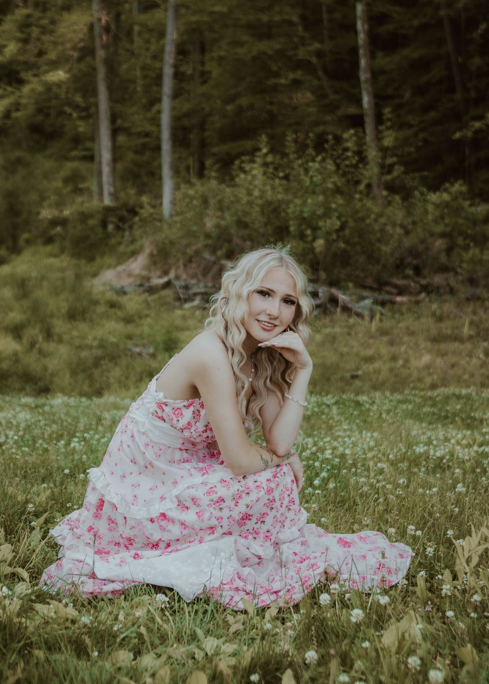 Blonde woman in pink floral dress sitting in wildflower meadow with forest background.