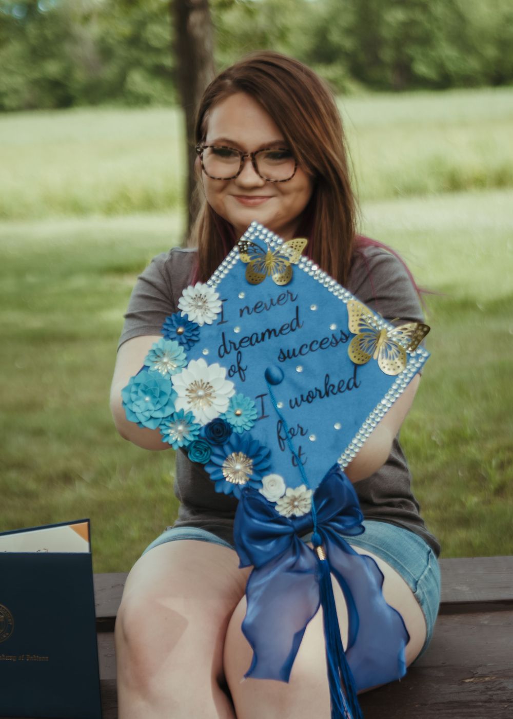 Graduate holds a decorated blue cap reading 'I never dreamed of success, I worked for it' with floral accents.