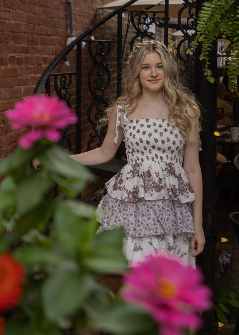 Blonde teenage girl in tiered floral dress posing on iron staircase surrounded by pink flowers.