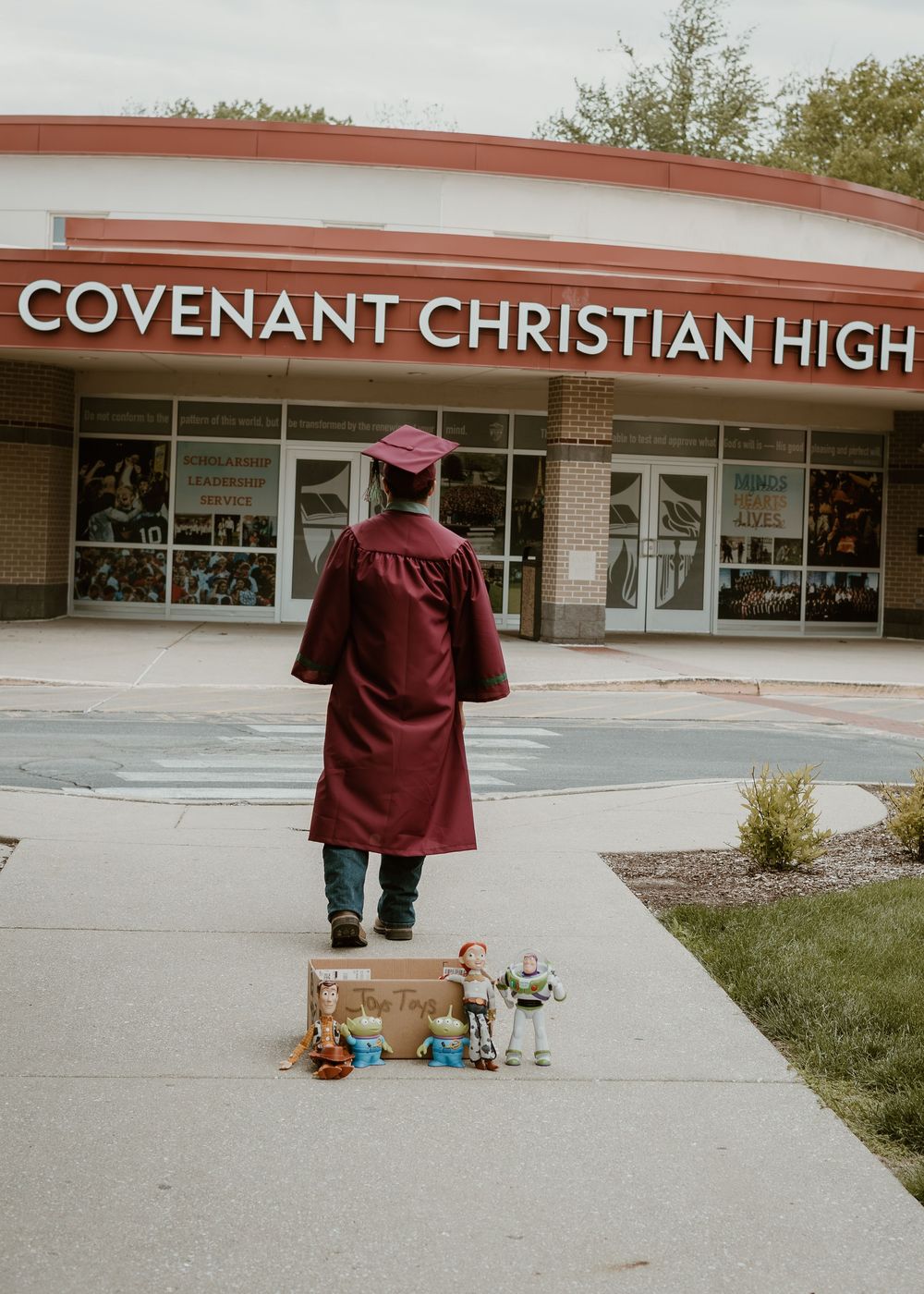 Graduate in maroon cap and gown standing before Covenant Christian High School with toys.