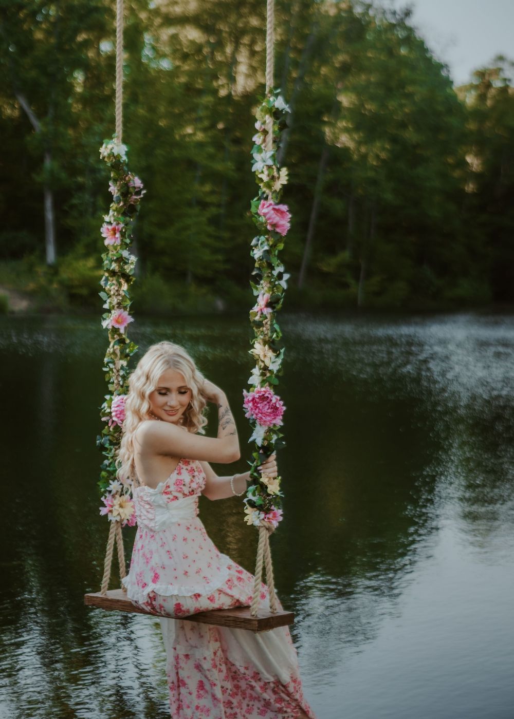 Blonde woman in a pink floral maxi dress sits on a flower-adorned swing over a reflective lake.