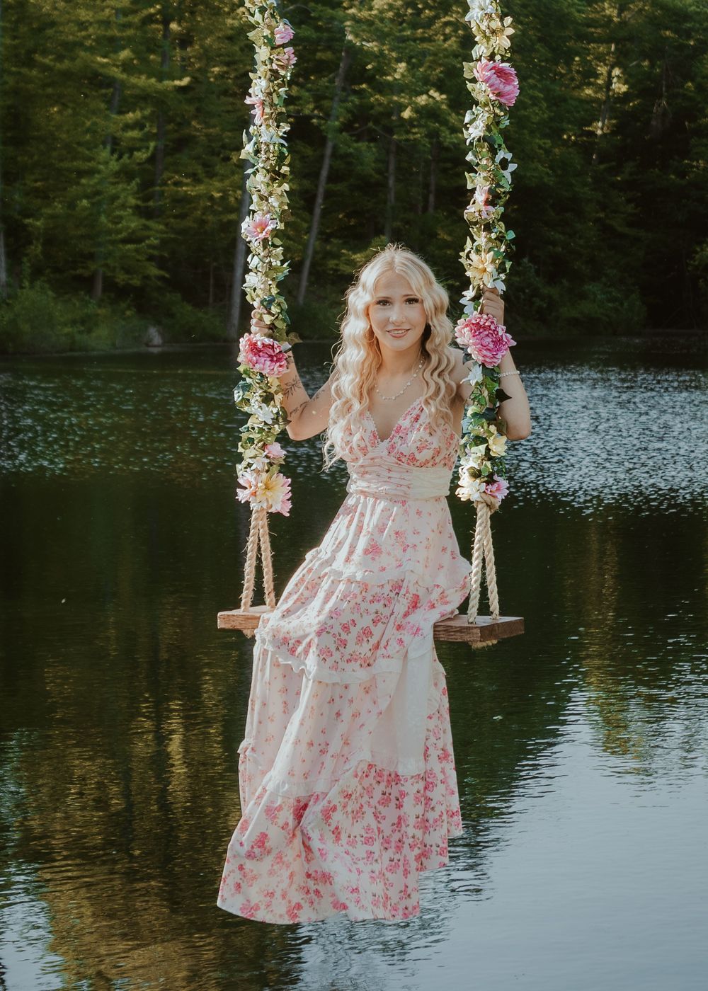 Smiling blonde woman in a pink floral dress sits on a flower-decorated swing above a serene lake.