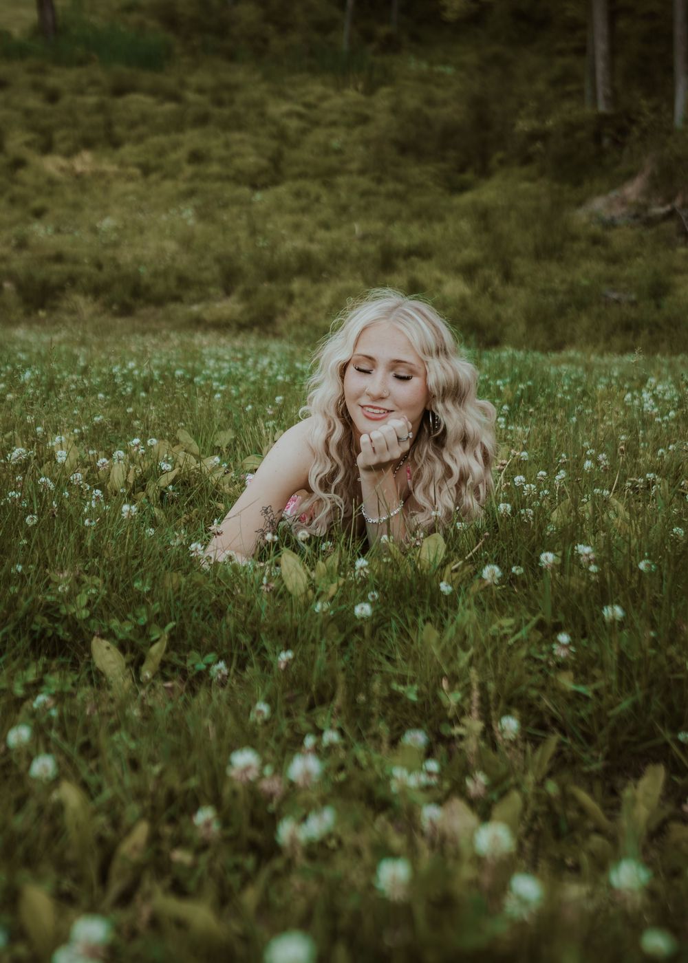 Blonde woman lies in a lush green meadow filled with small white wildflowers, smiling softly.