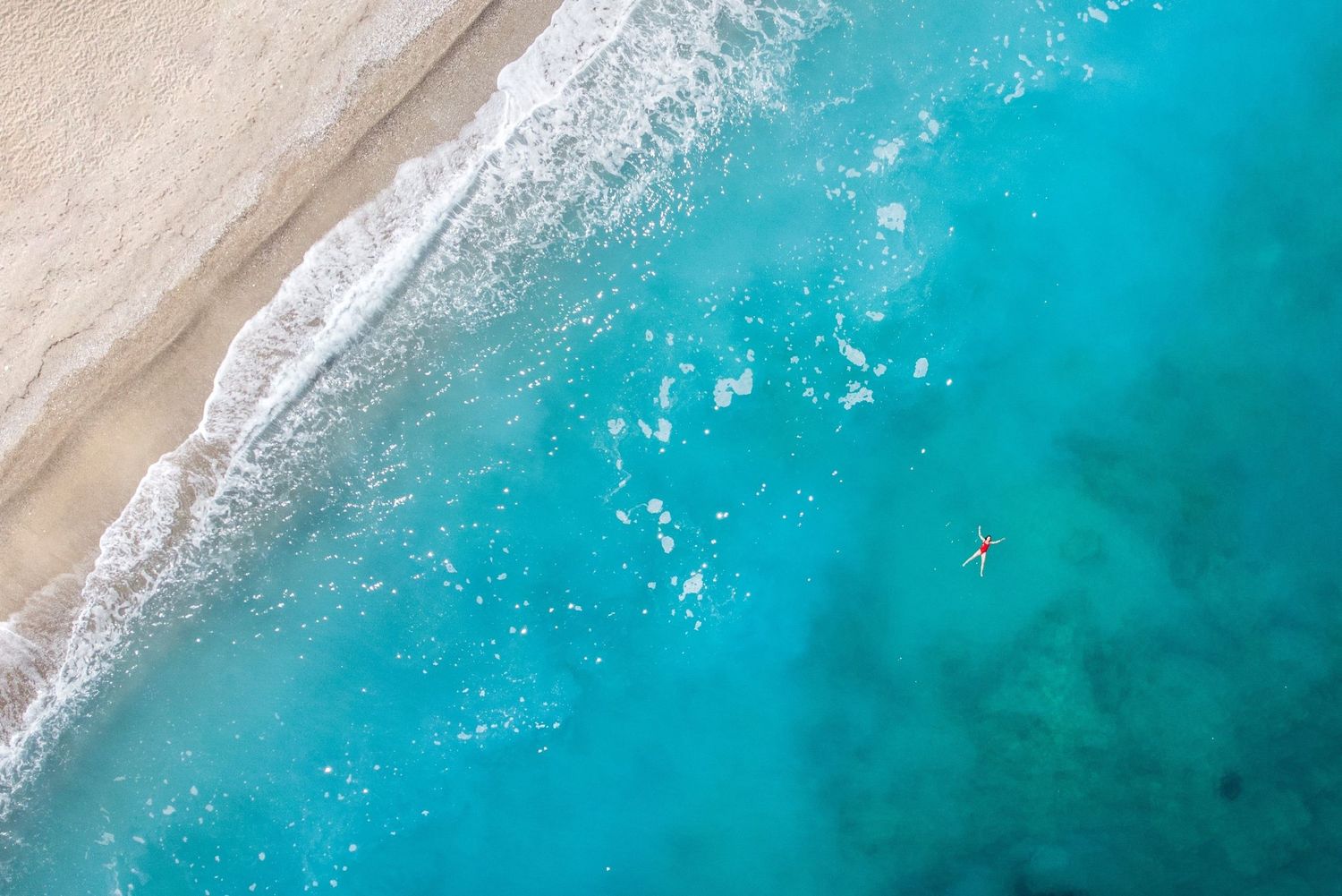Aerial view of a lone swimmer in crystal clear turquoise ocean water beside a sandy beach with gentle waves.