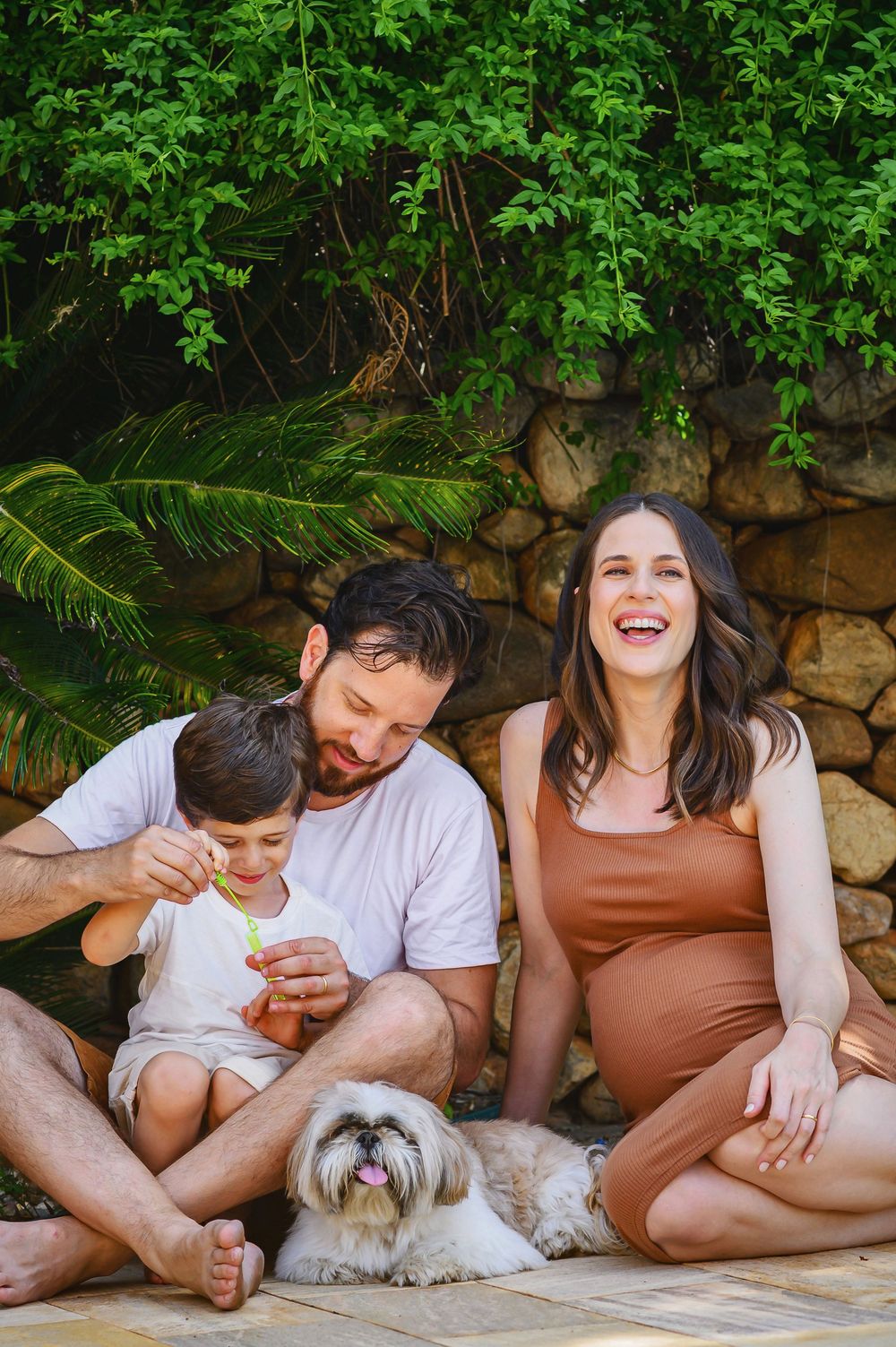 Happy family with a young boy, pregnant woman, and man sitting outdoors with a fluffy dog near a stone wall.