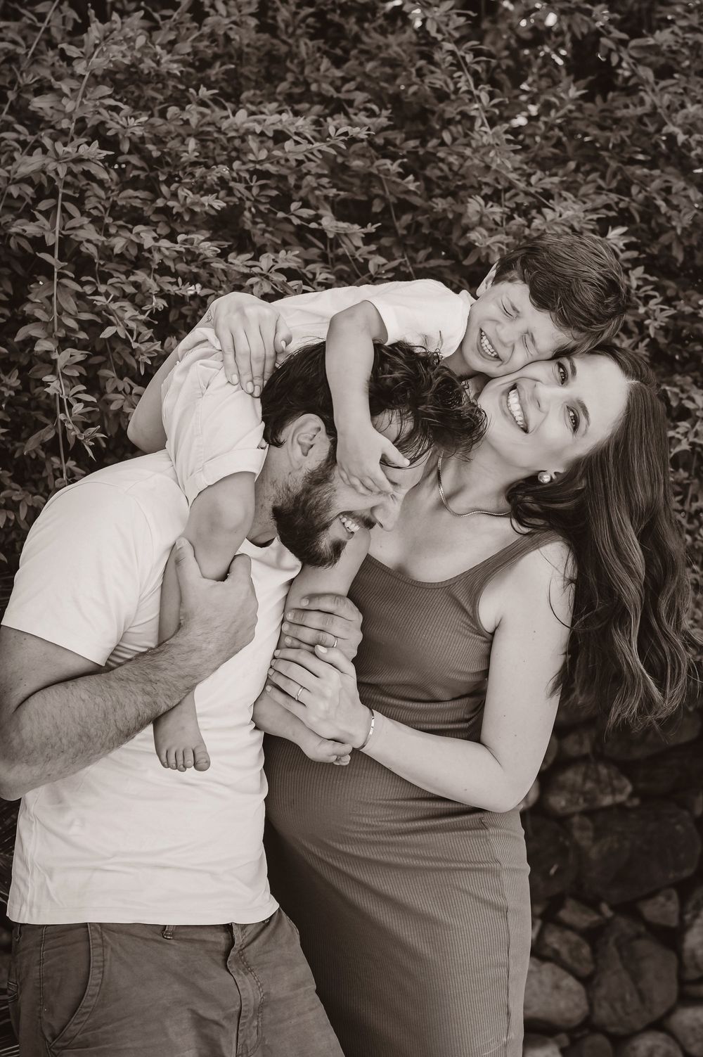 Black and white photo of a happy pregnant woman and man playfully hugging a child outdoors near lush greenery.