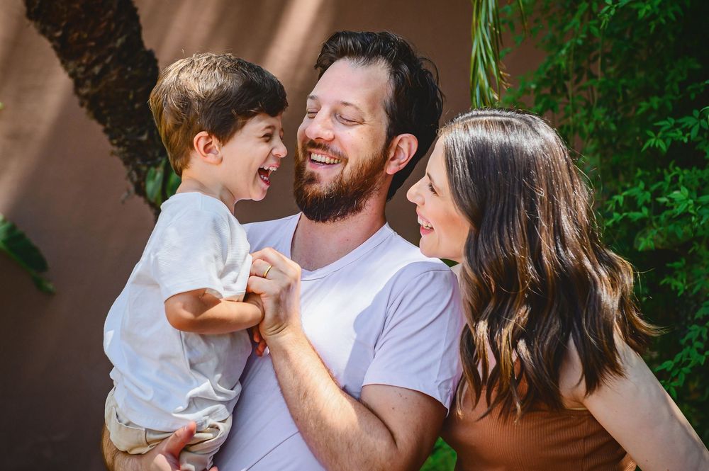 Happy family portrait with father holding young son while mother smiles, outdoors with greenery background.