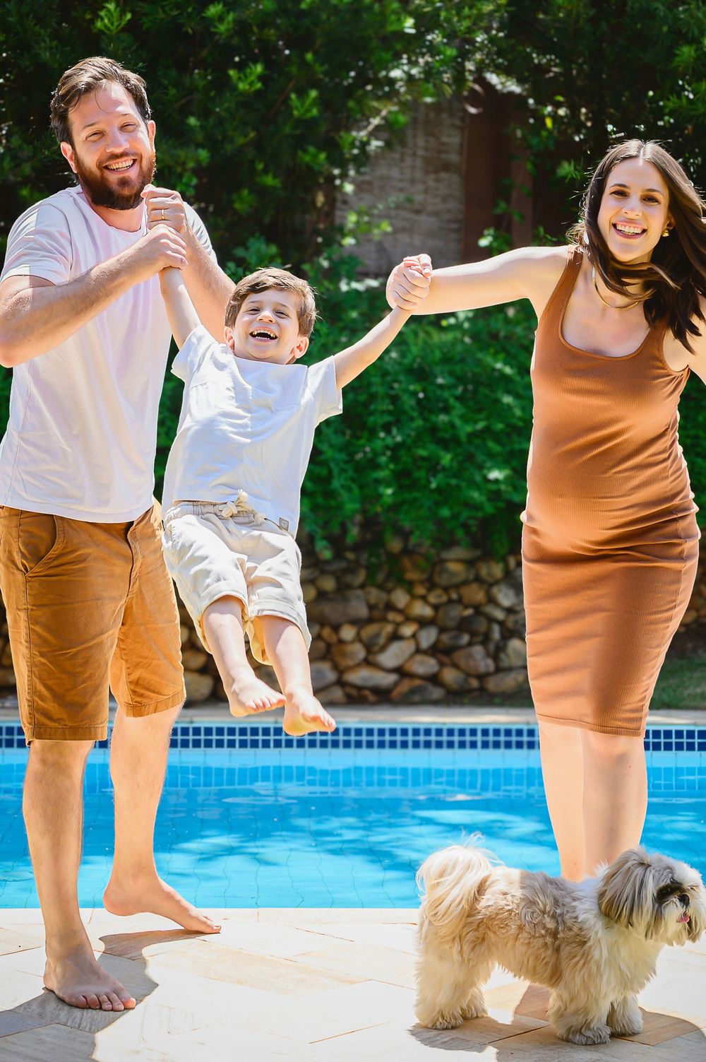 Happy family with parents swinging child by the pool, with a fluffy dog nearby on a sunny day.