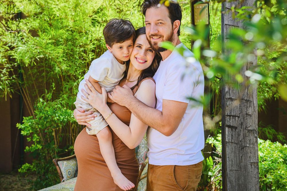 Happy family of three posing outdoors, pregnant mother holding toddler son, father embracing them in lush green garden.