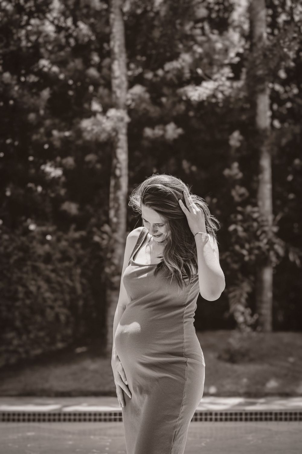 Pregnant woman in a fitted dress smiling outdoors near a pool, black and white maternity photo.