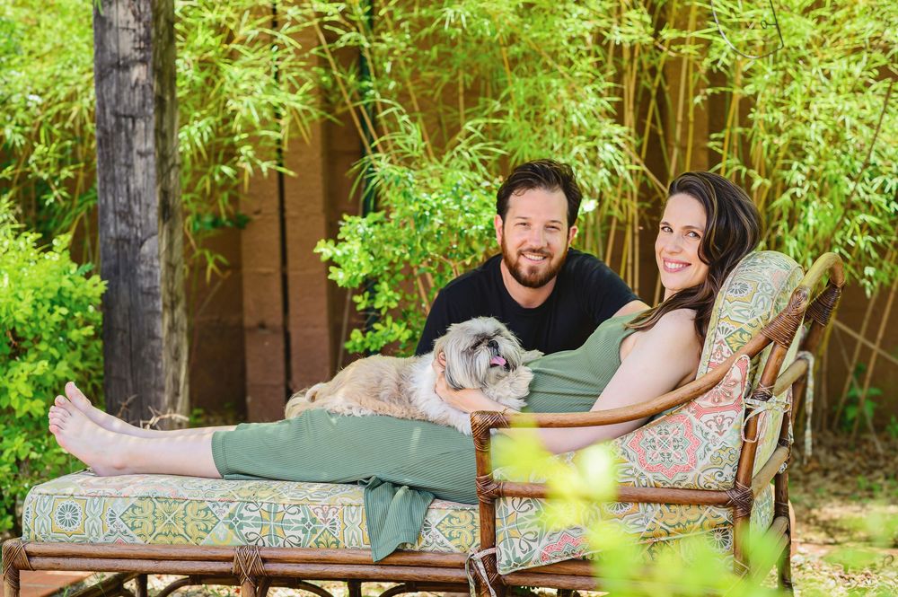 Pregnant couple relaxing outdoors on a lounge chair with their small gray dog, surrounded by lush green bamboo.