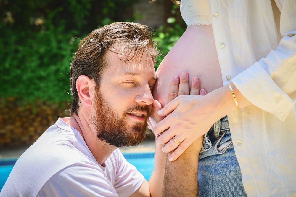 Bearded man kneeling and pressing his cheek against a pregnant woman's bare belly outdoors near a pool.