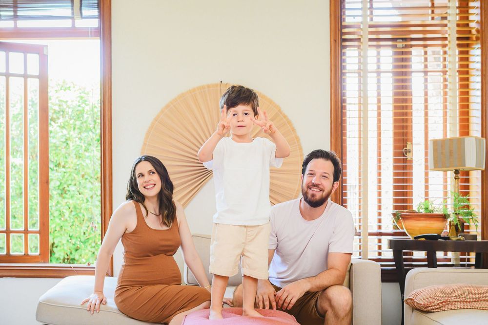 Happy family of three posing indoors, pregnant mother in brown dress, father smiling, young boy making peace signs.