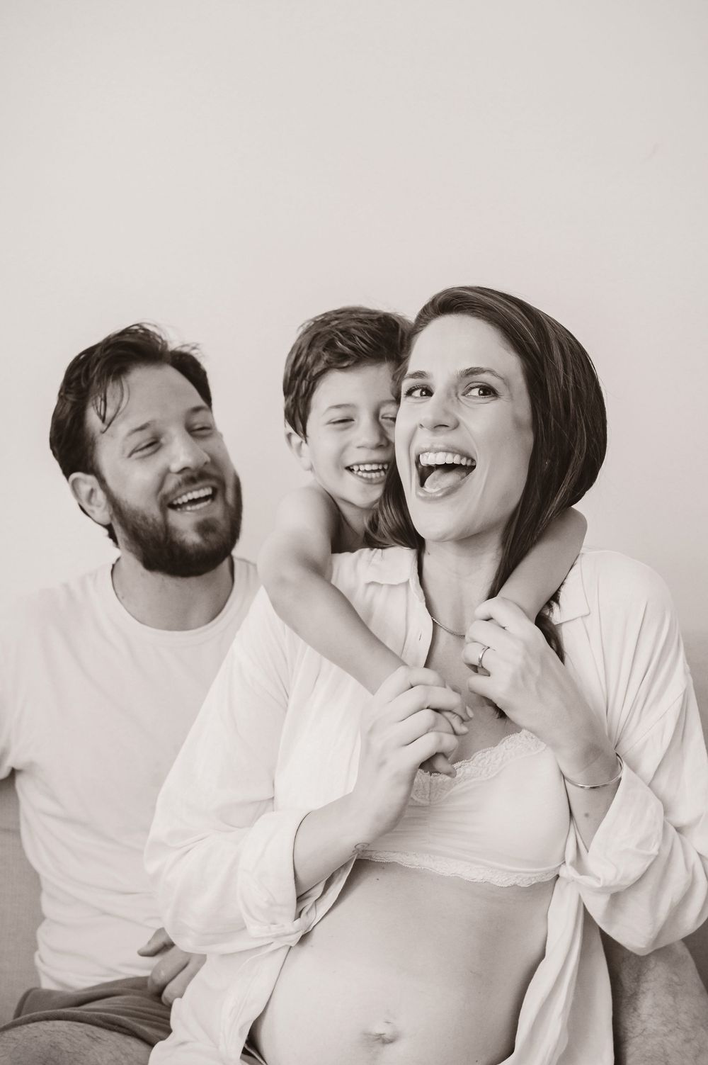 Black and white maternity photo of laughing pregnant woman with bearded man and young boy hugging her from behind.