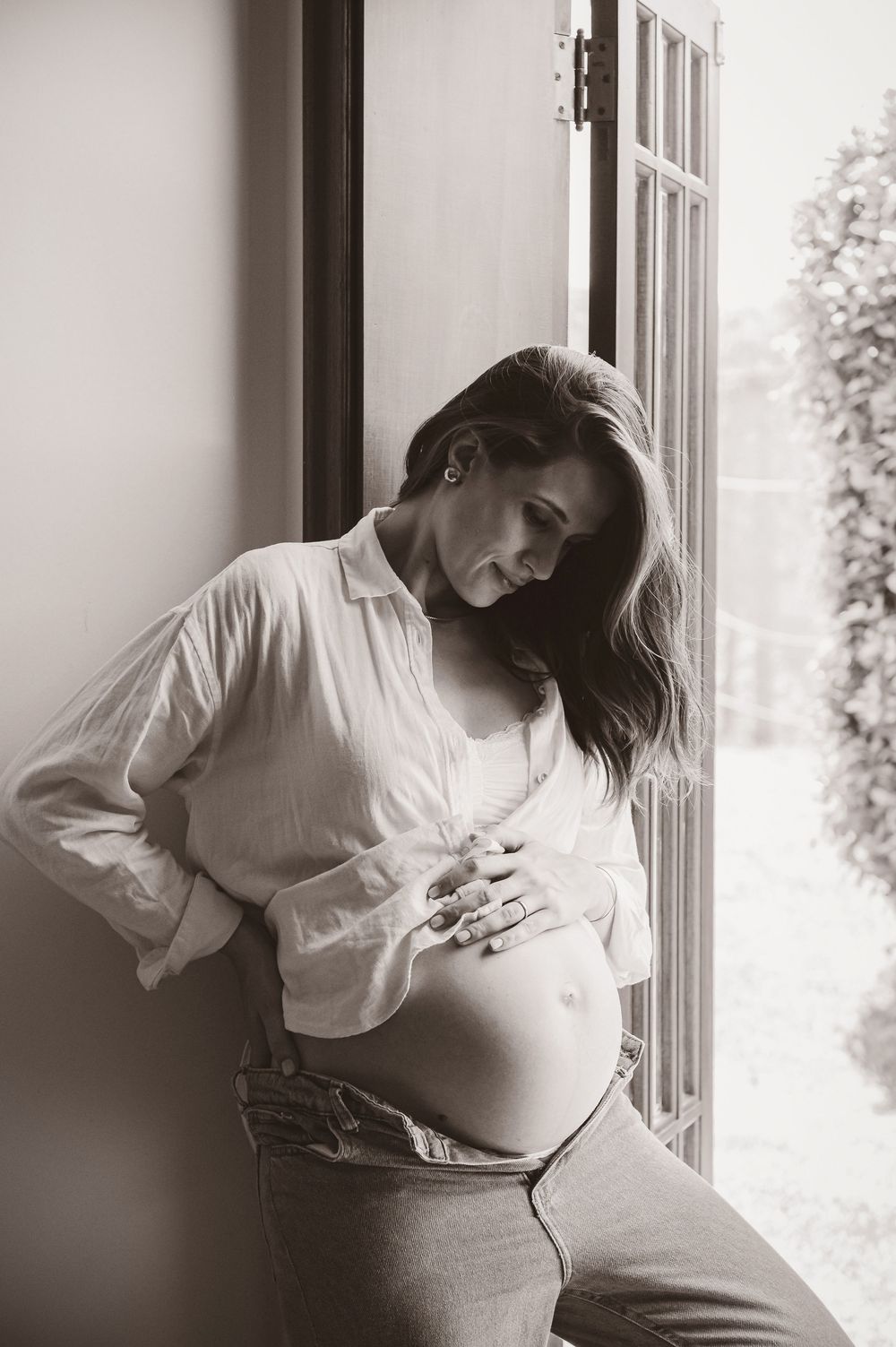 Black and white photo of a pregnant woman cradling her baby bump while leaning against a window.