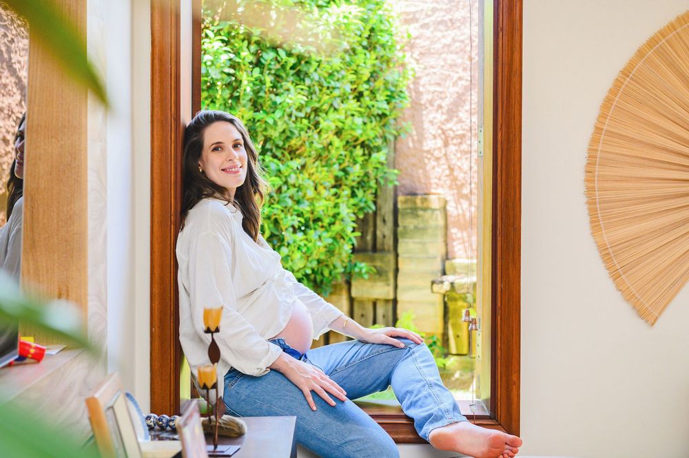 Woman in white blouse and jeans sitting by open glass door with lush green wall backdrop in cozy home interior.