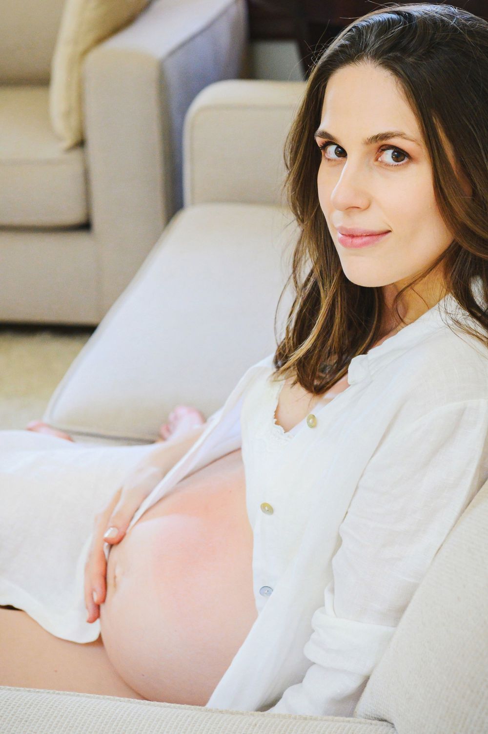 Pregnant woman in white shirt sitting on floor leaning against sofa, cradling bare baby bump at home.