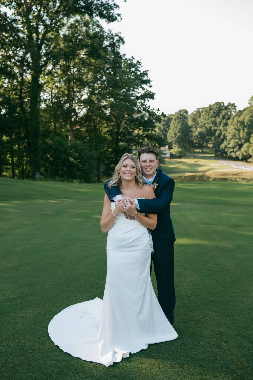 Bride and groom embrace on a lush golf course, she in a white gown, he in a navy suit, surrounded by trees.