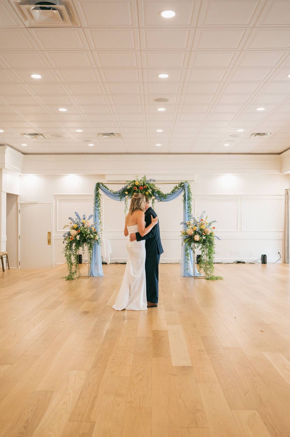 Bride and groom share first dance in elegant ballroom with floral arch backdrop and blue draping.
