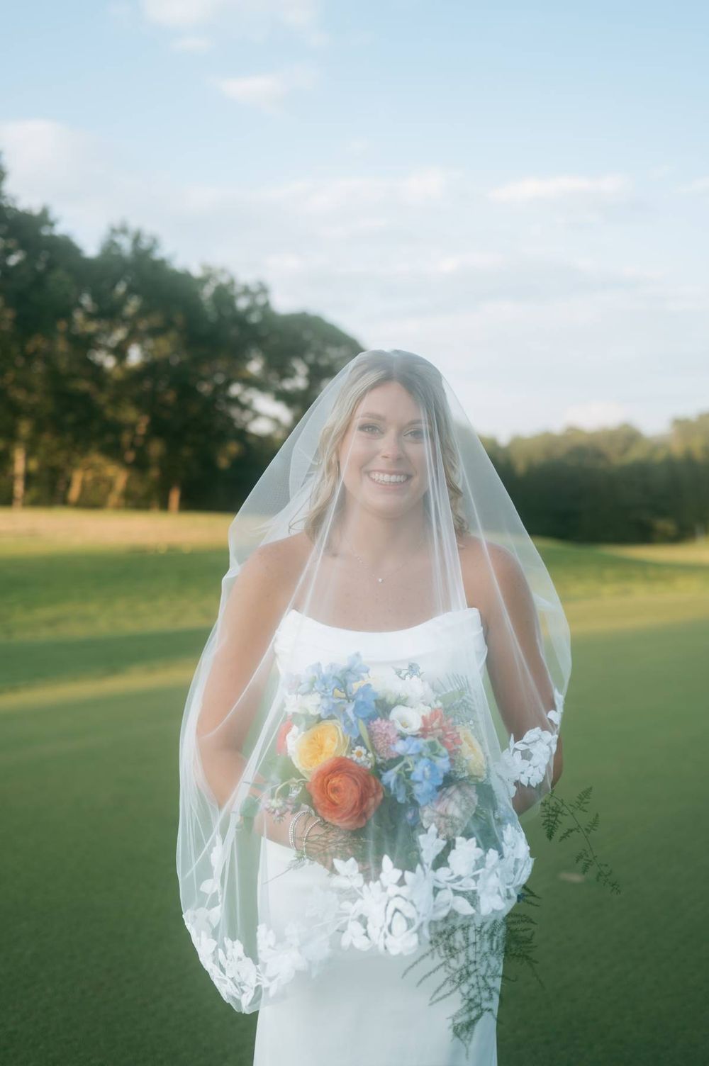 Smiling bride in white gown and lace veil holds colorful bouquet outdoors on green lawn with trees.